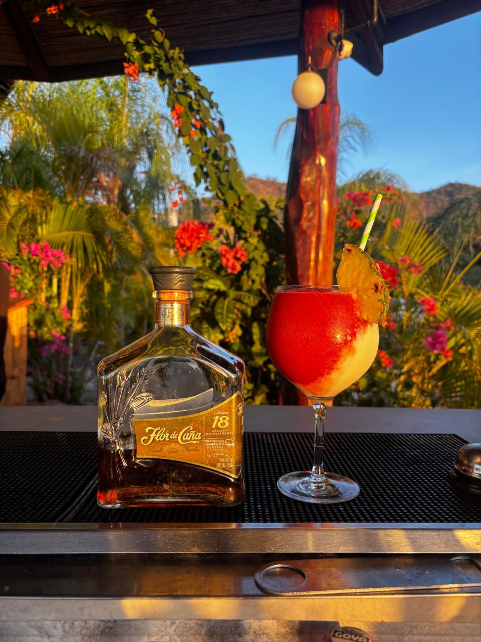 Botella de ron Flor de Caña 18 junto a un daiquirí de fresa y piña al atardecer